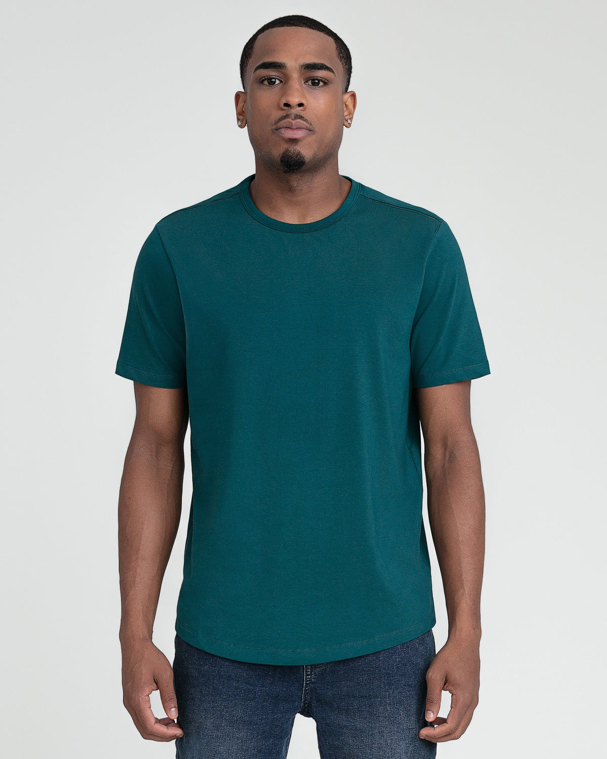 Man wearing teal crew neck short-sleeve t-shirt with dark jeans against gray background.