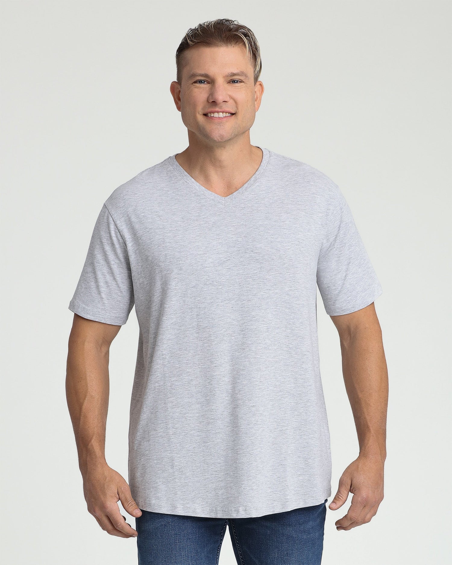 Man wearing light gray V-neck t-shirt with short sleeves and dark jeans against white background.