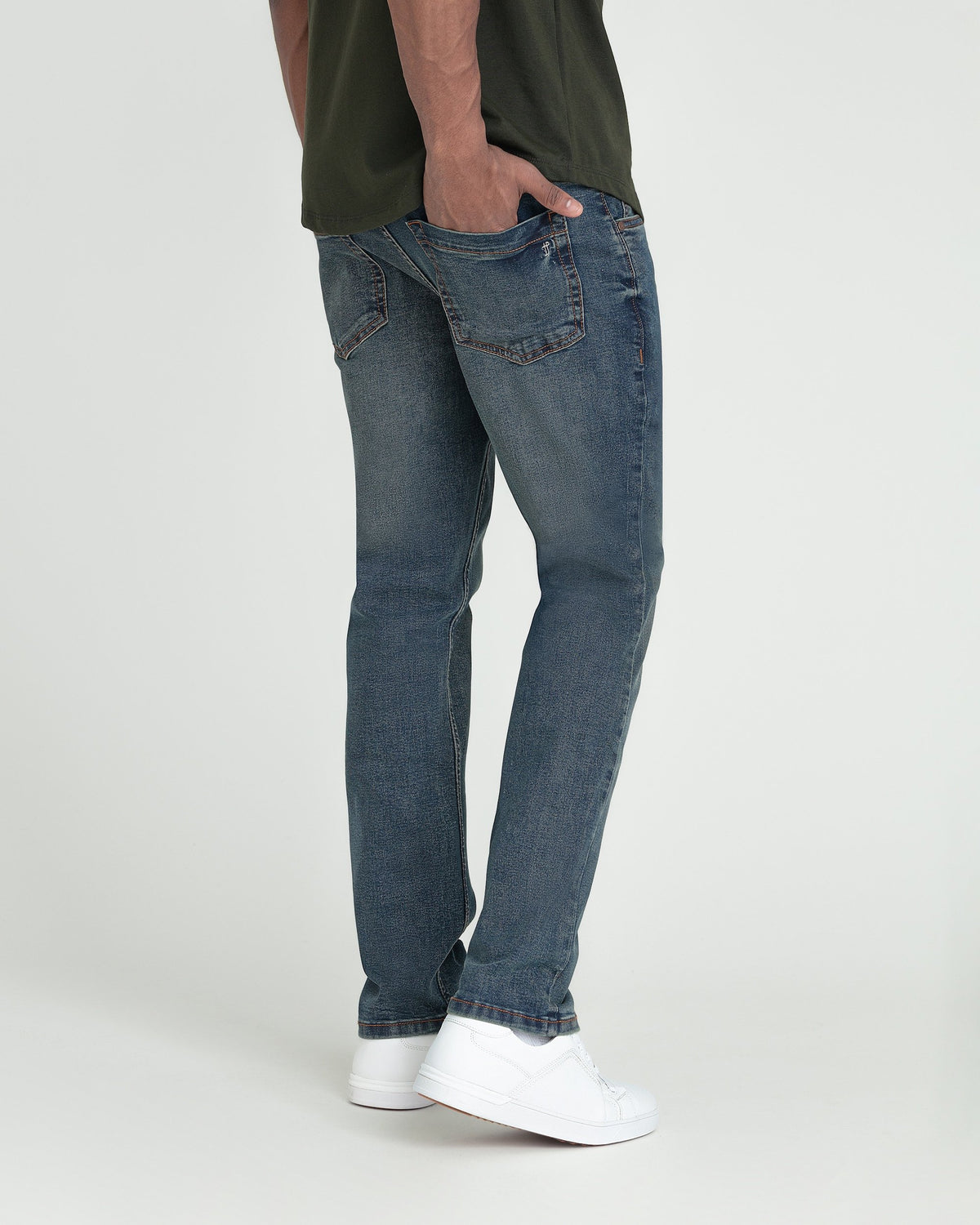 Slim fit medium-wash denim jeans with straight leg, worn with olive shirt and white sneakers.