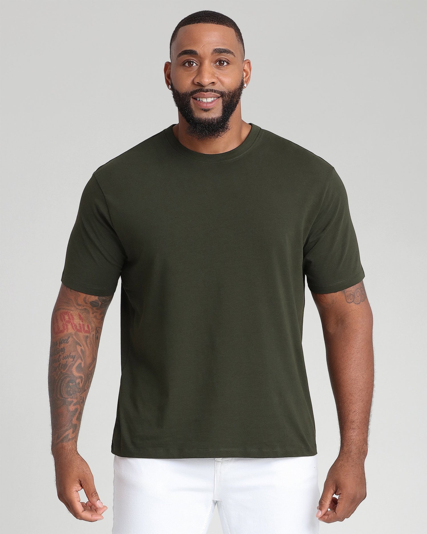 Man wearing olive green crew neck t-shirt with white pants against gray background.