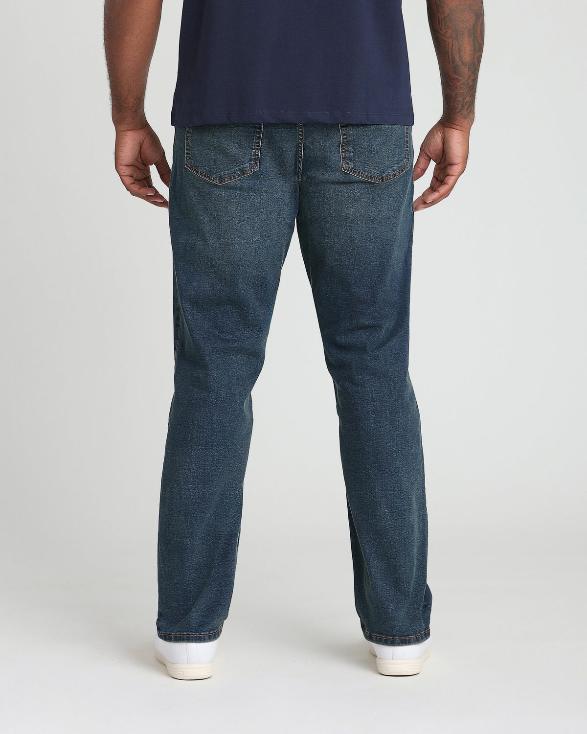 Man wearing dark blue t-shirt and tinted blue denim jeans with white sneakers, back view.