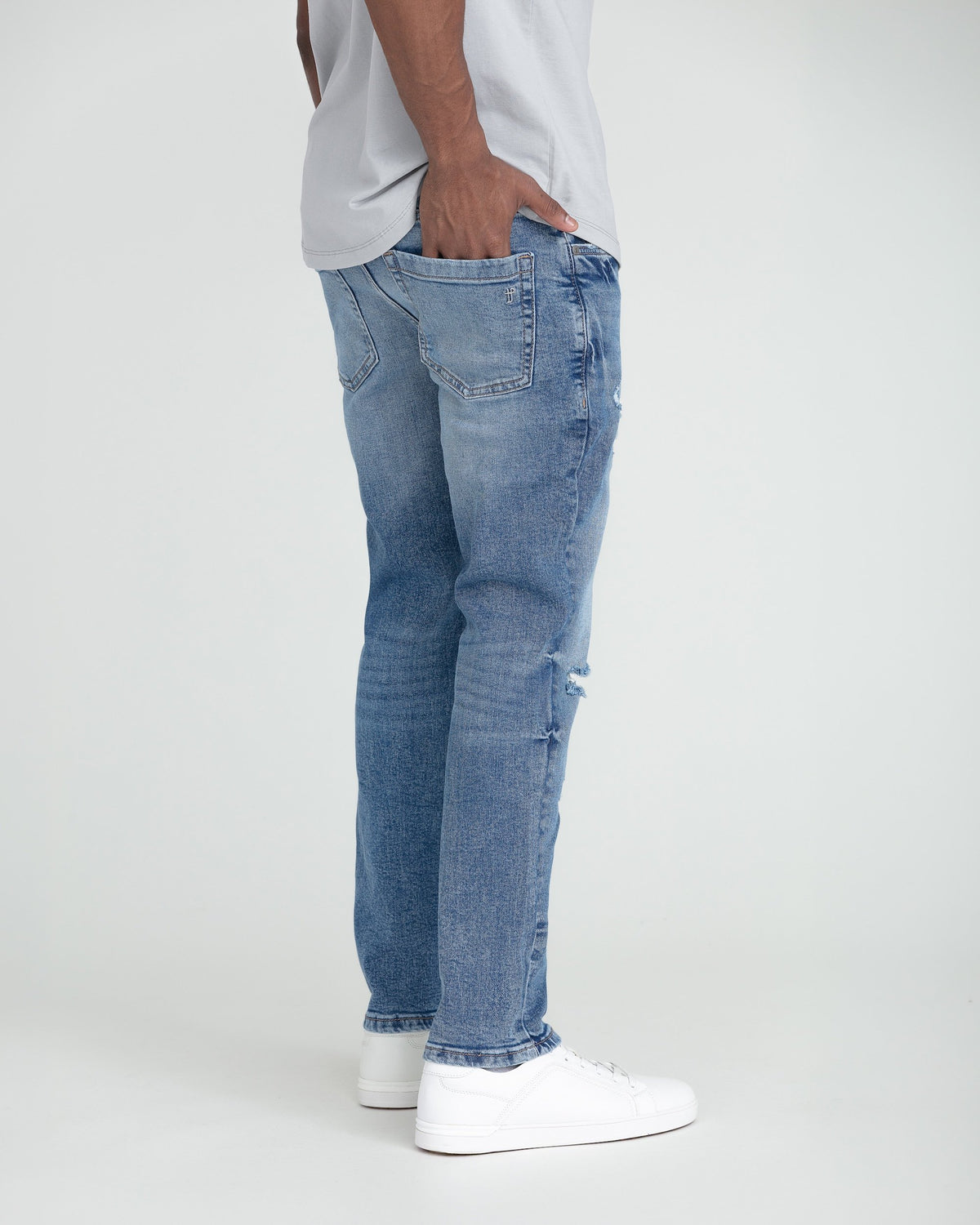 Medium blue slim fit jeans with ripped detail on thighs, shown on model wearing white sneakers.