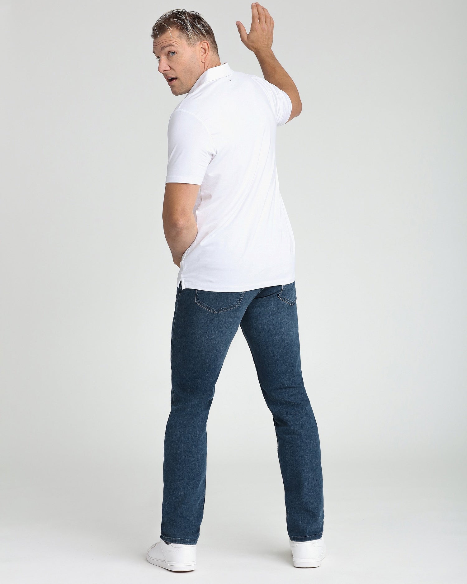 Man wearing white polo shirt with dark blue jeans and white sneakers, shown from behind in three-quarter view.